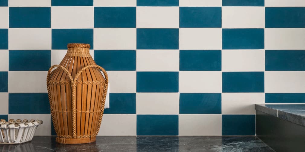 A decorative wicker vase sits on a dark marble surface in front of a blue and white checkerboard tile wall. A small dish with round objects is also visible on the left side of the image.